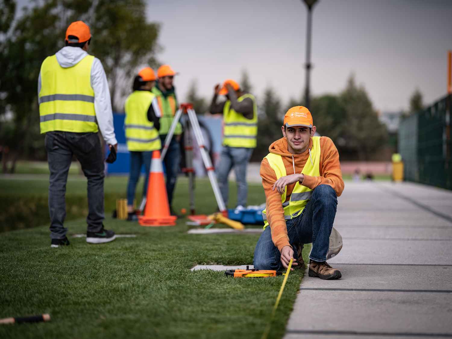 Estudiante de ingeniería mide distancia con cinta métrica sobre una banqueta mientras compañeros usan equipo de topografía al fondo.